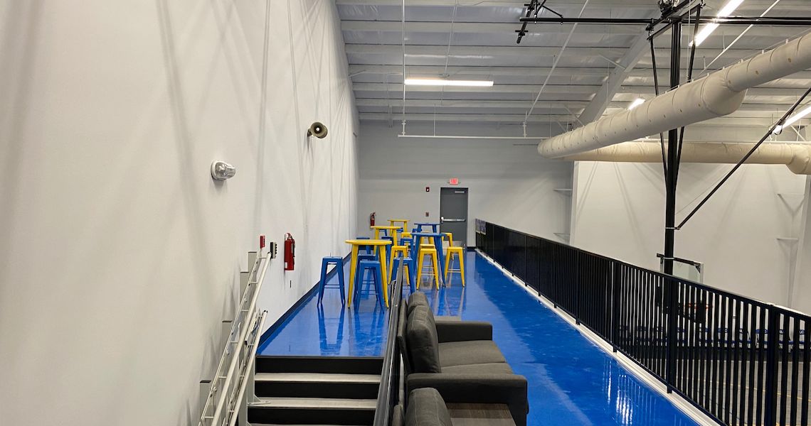 A modern indoor space reminiscent of a charter school, featuring a vibrant blue floor with several yellow and blue bar stools lined up against a wall. A row of gray couches graces the opposite side, complemented by black railings, while the ceiling showcases exposed pipes and lights.