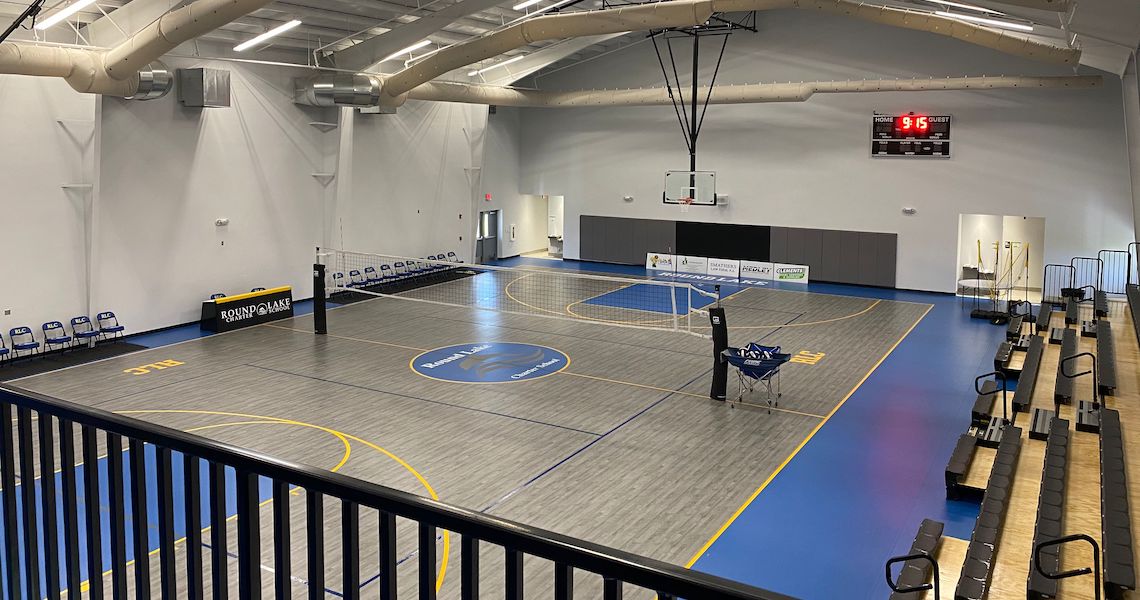 An empty indoor basketball court at Round Lake Charter School boasts a polished wooden floor. Volleyball nets are set up, and bleachers line the side. The scoreboard shows 9-5. Exposed beams and ductwork adorn the ceiling, while chairs neatly line one wall.