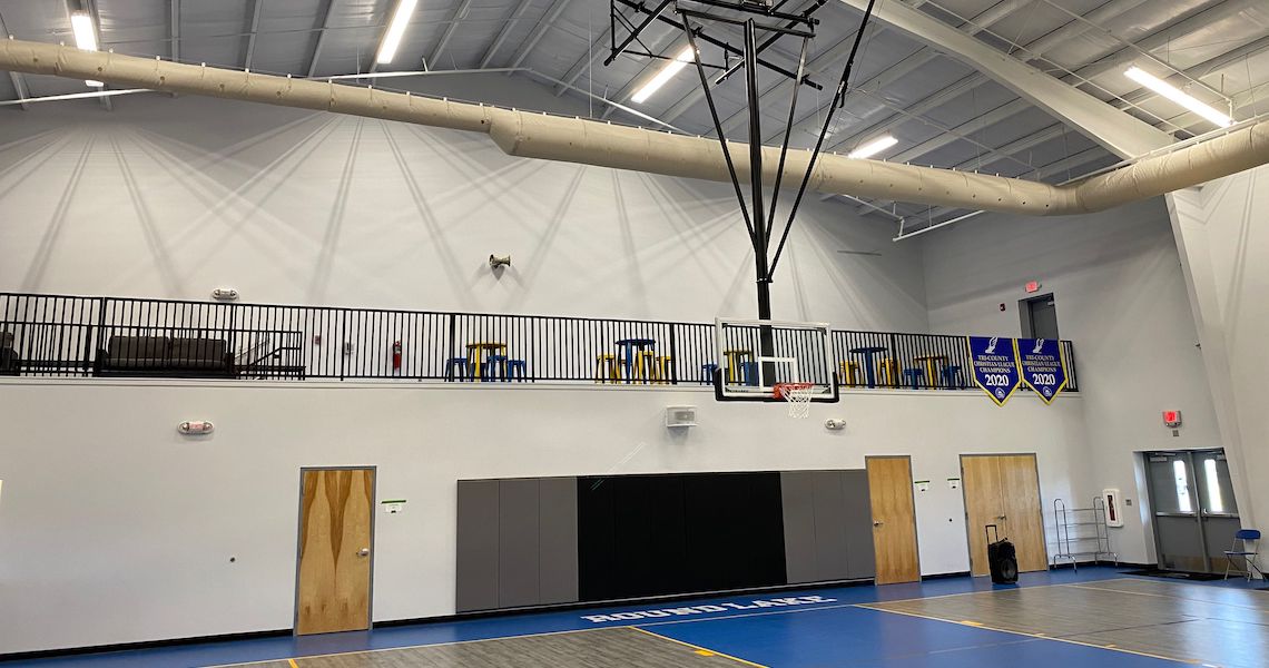 The gymnasium at Round Lake Charter School features a basketball hoop, blue and gray flooring, and a second-level seating area with chairs. Championship banners hang from the railing, adding pride to the scene. Two wooden doors and a wall panel are noticeable on the side.