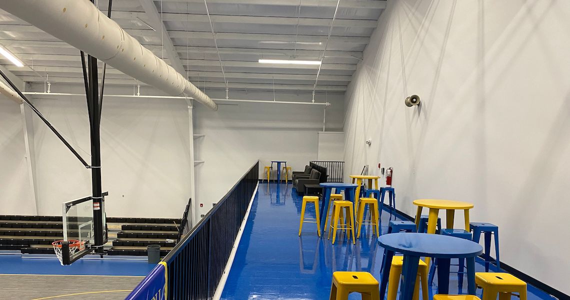 Elevated indoor sports area at Round Lake Charter School features a blue floor with yellow and blue stools and tables. It overlooks a basketball court with bleachers, framed by white walls and ceiling, complete with visible air ducts and lighting.