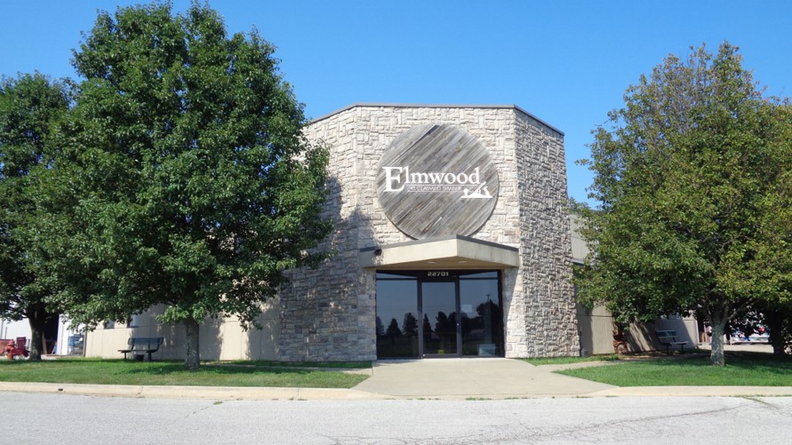 A building with stone and beige walls, featuring a round wooden accent above glass entrance doors. The sign reads "Elmwood." It is surrounded by two leafy trees and sits under a clear blue sky.