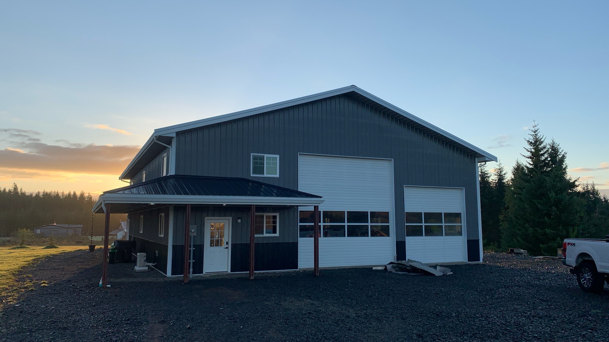 A large, gray metal building with two garage doors is set against a backdrop of trees at sunset. The building has a small porch with a door and a window above. A white vehicle is parked nearby on a gravel surface.