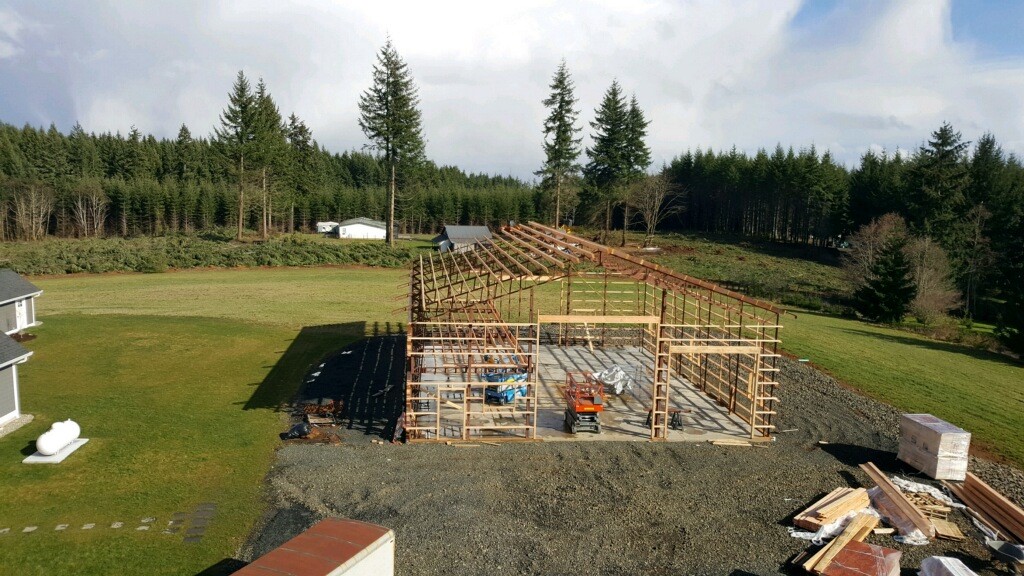 Aerial view of a wooden barn under construction in a rural area, surrounded by green fields and tall pine trees. Building materials and equipment are scattered around the site. A white house is visible nearby.