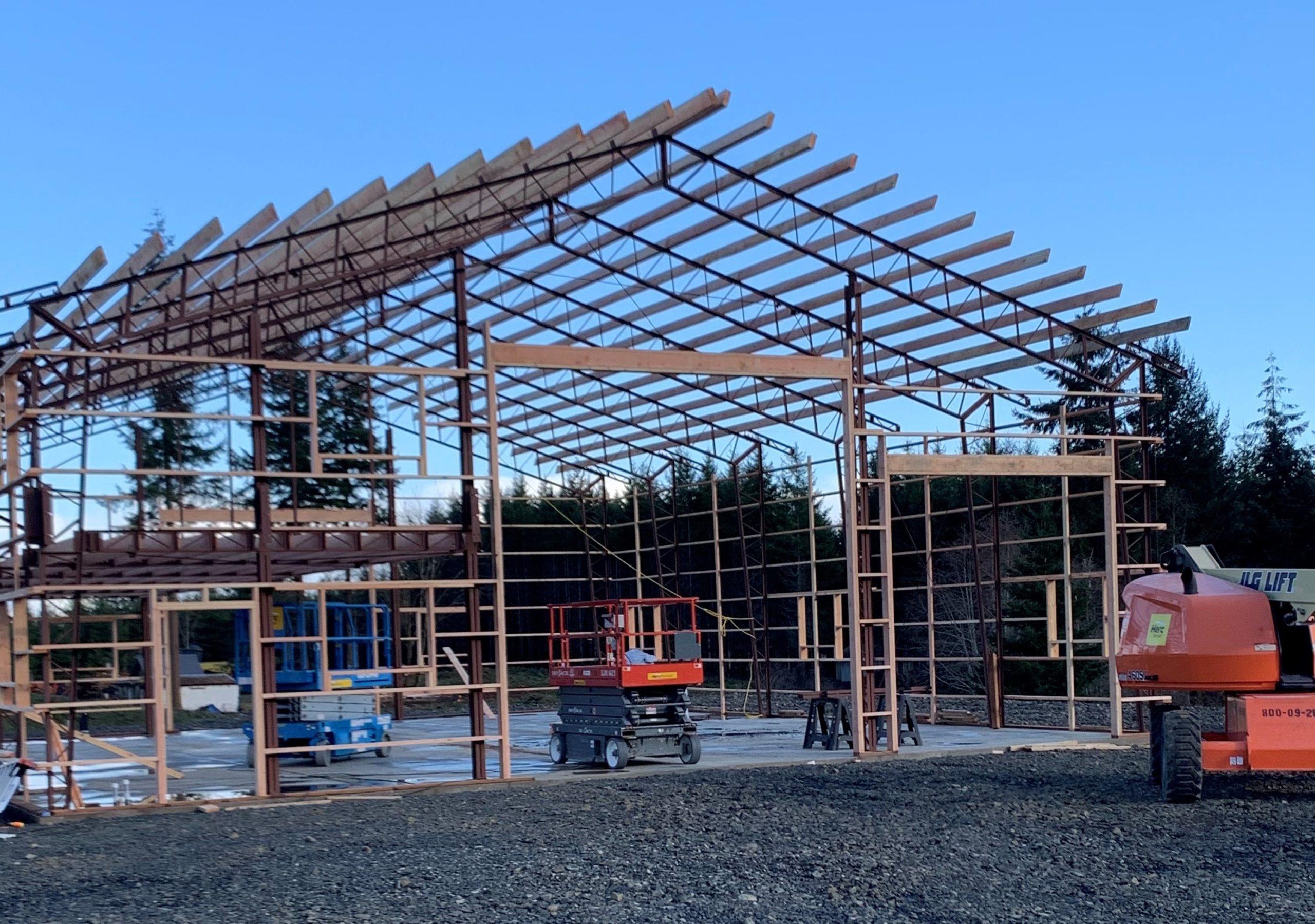 A large metal framework of a building under construction, with visible beams and supports. Various construction equipment, including a blue lift and an orange forklift, are on site. The background shows trees under a clear blue sky.