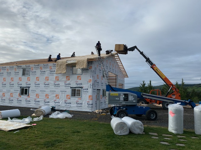 A house under construction with workers installing roof insulation. The building is wrapped in Tyvek sheeting. A forklift lifts materials to the roof, and various tools and insulation rolls are on the ground. Overcast sky in the background.