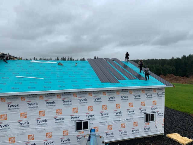 Two workers are installing metal roofing sheets on a house under construction. The house is wrapped with Tyvek, and the sky is overcast. The surroundings are green with grass and trees.