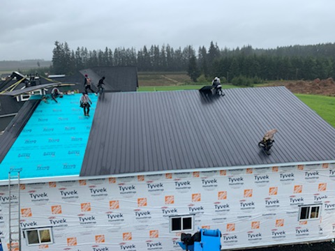 Workers install a metal roof on a large building amid a forested landscape. Part of the roof is completed, while some workers position materials on a turquoise underlayment. Overcast skies and machinery are visible in the background.