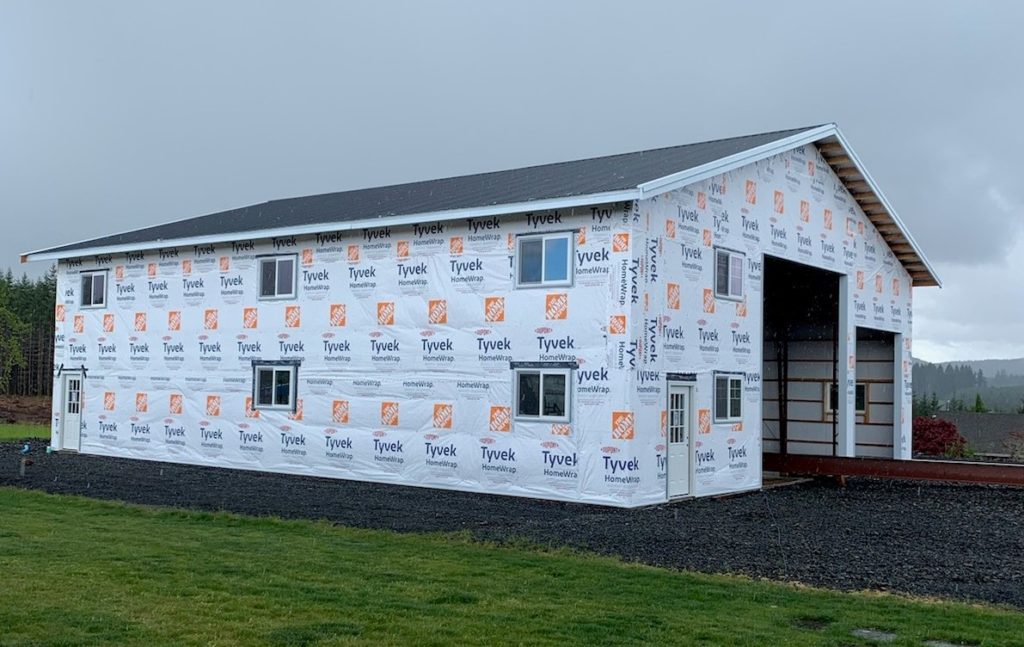A large barn-style building under construction with a wrap labeled "Tyvek HomeWrap" covering its exterior. The building is situated on a gravel area with green grass in the foreground and an overcast sky in the background.