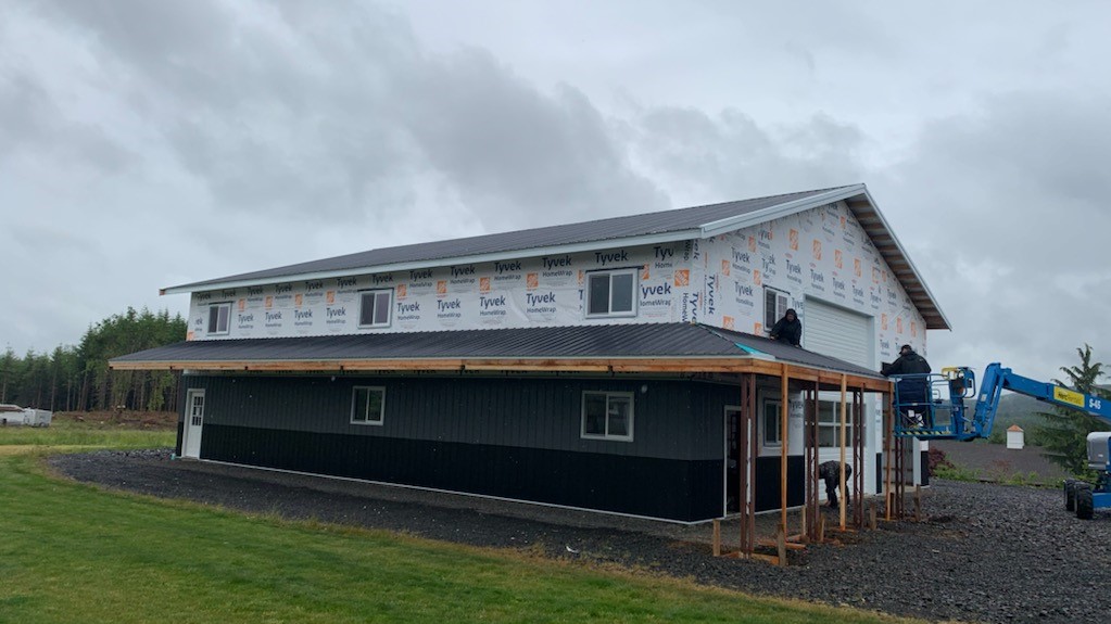 A large building under construction with a partially completed roof and walls covered in Tyvek wrap. Workers are on a lift and the ground, and a grassy area surrounds the construction site. Overcast sky in the background.