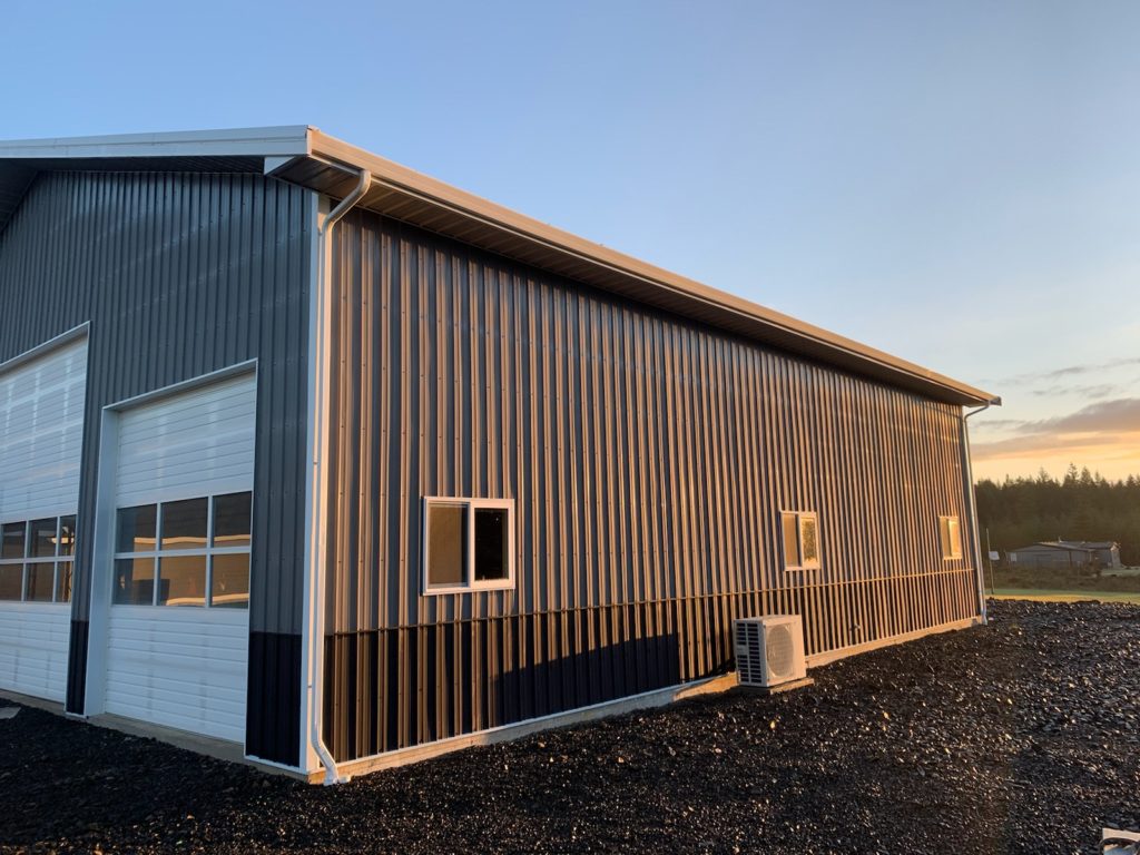 A large modern metal building stands under a clear blue sky. The structure features vertical siding and several small windows. The ground is covered in gravel, and the sun casts a warm glow on the side of the building. A rural landscape is visible.