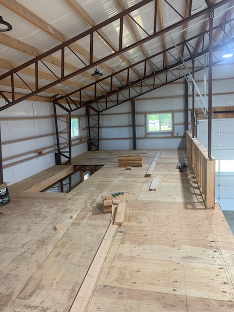 Interior of a partially constructed building with wooden flooring. The space has metal beams and a high ceiling. Construction materials, tools, and windows are visible, allowing natural light to illuminate the area.