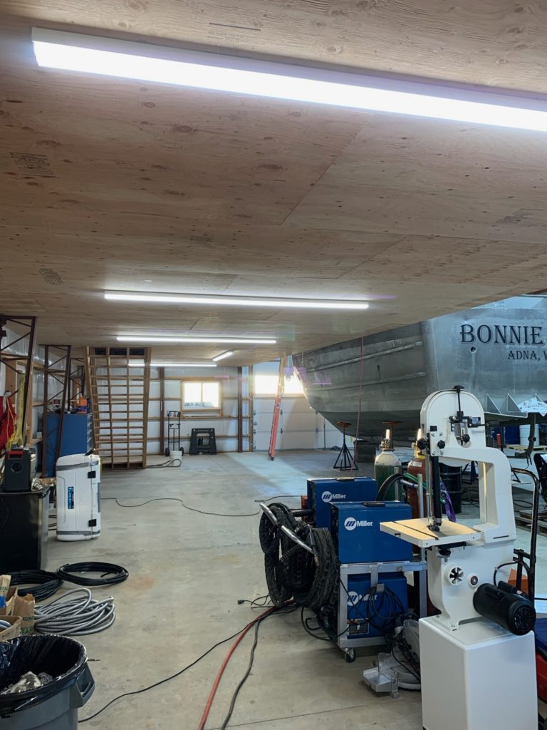 A spacious garage workshop with bright overhead lights. A metal boat labeled "BONNIE" is on the right, with various tools and equipment, including a blue welder and a white machine, scattered around the concrete floor.