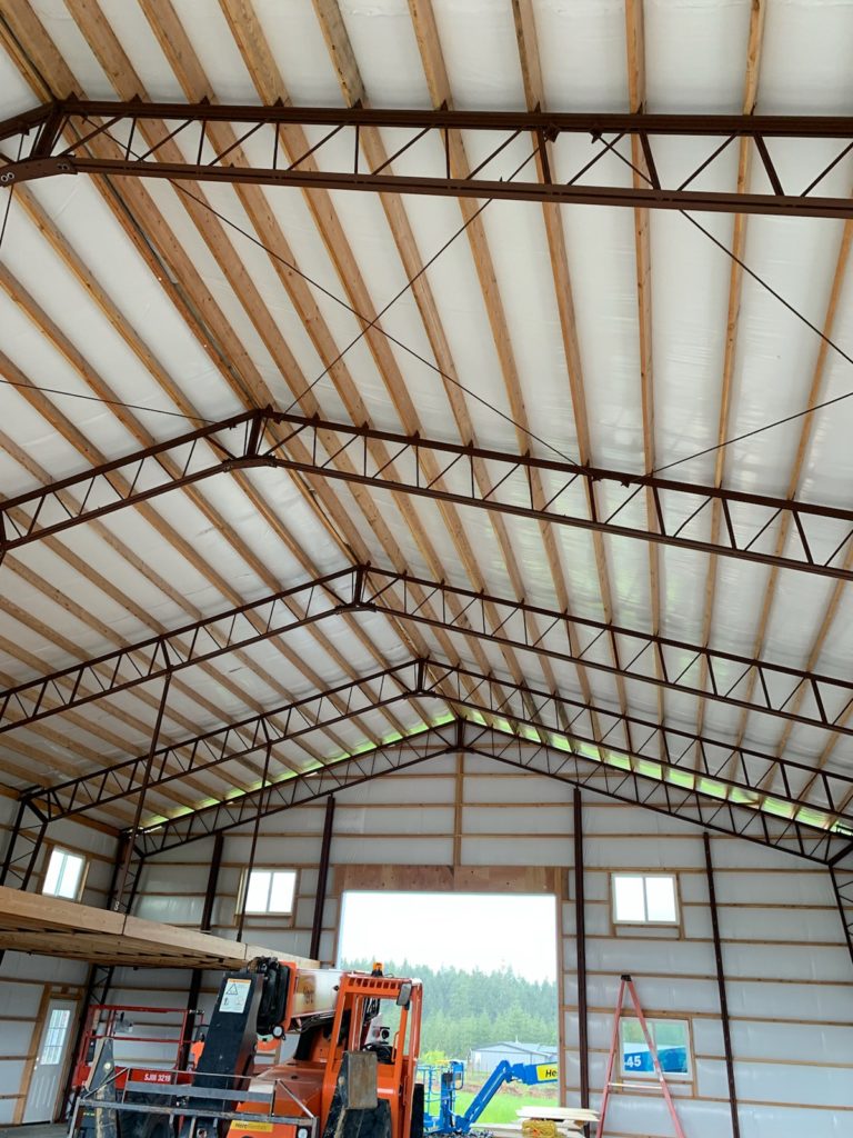 Interior view of a large warehouse or barn with metal and wooden beams, white insulation on the ceiling, and construction equipment, including a lift and scaffolding, on the floor. Small windows provide natural light.