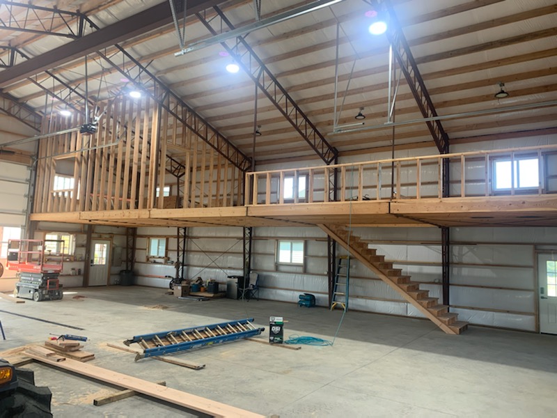 A spacious warehouse interior with a high ceiling, featuring a wooden mezzanine and staircase on the right. Construction materials and tools are scattered on the concrete floor, including a ladder, saw, and planks. Natural light enters through windows.