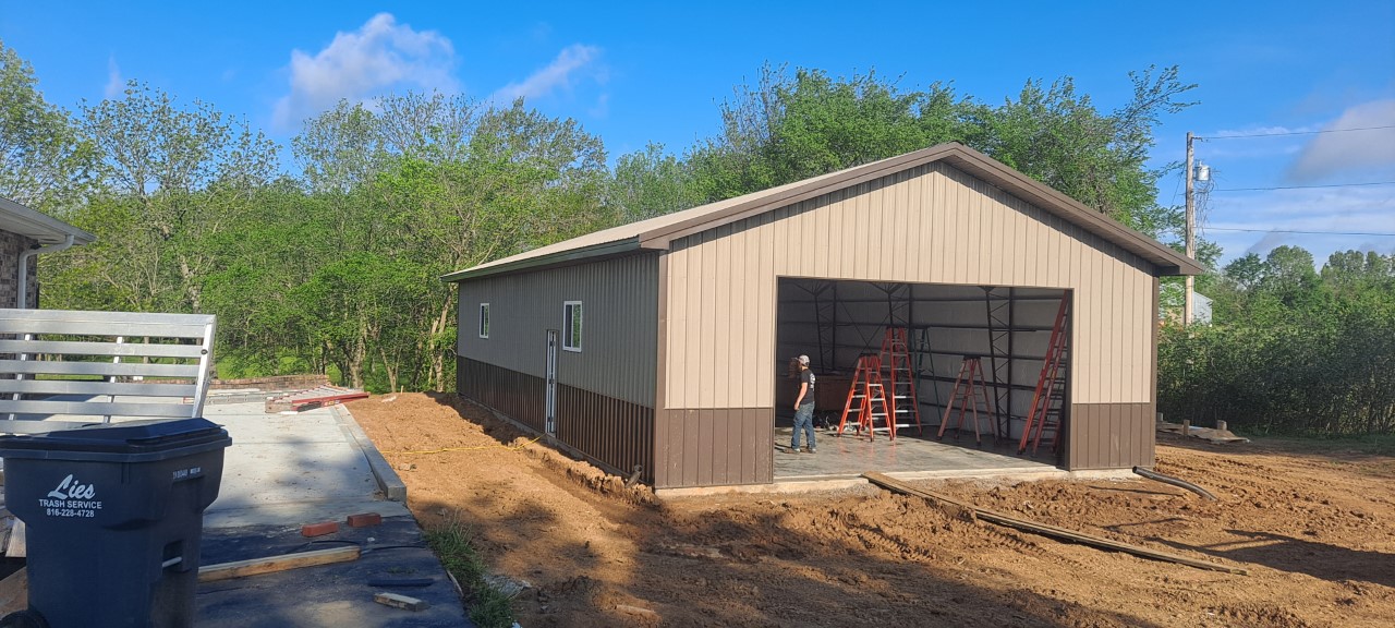 A large, unfinished garage with a brown and beige exterior stands on a dirt area in Grain Valley. Several ladders are set up at the entrance. A person is working inside the shop. Trees and a blue sky fill the background.