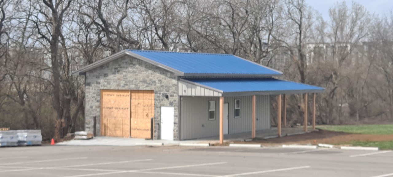 A small building with a blue metal roof and stone facade sits next to an empty parking lot, resembling an old Fraternal Order of Police lodge. The building has a porch with wooden posts and boarded-up windows, while leafless trees stand silently in the background.