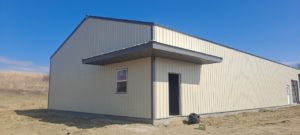 A beige metal building with a sloped roof and a small awning over the entrance serves as a dog training facility. Set amid dry grass under a clear blue sky, it features a single window and an open door on the front side, welcoming canine companions and their owners.