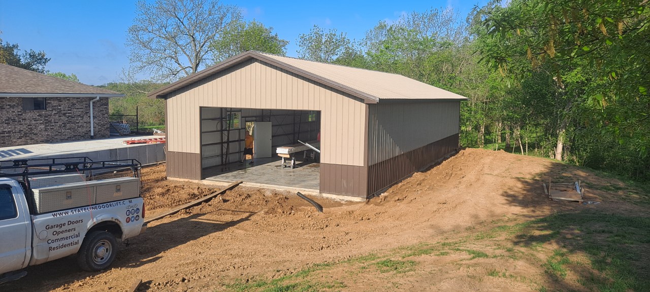 A partially constructed building in Grain Valley, featuring an open garage door, stands on a dirt foundation surrounded by trees. A truck with company contact information is parked nearby on the left. The sky is clear and blue.