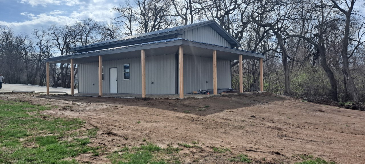 A small, gray metal building with a sloped roof and wooden support beams on the porch serves as the local Fraternal Order of Police lodge. It's situated on a dirt lot with bare trees in the background and a grassy area in the foreground, under a partly cloudy sky.