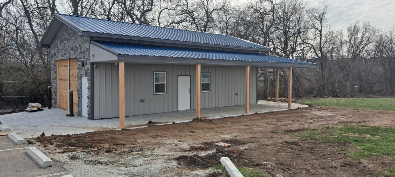 A small building, marked as an FOP lodge, with gray walls and a blue metal roof stands on a concrete foundation. The structure features wood pillars supporting an overhang. Surrounding the area are bare trees and a grassy field. A parking space is visible in the foreground.