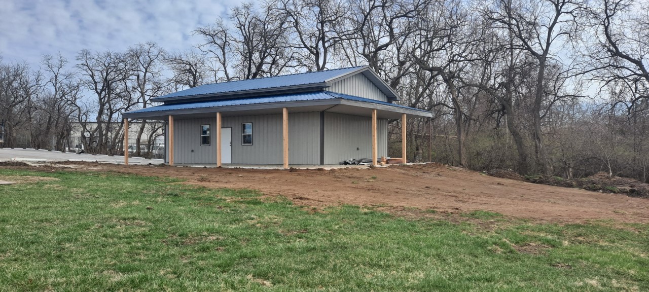 A small, gray building with a blue metal roof and wooden support beams, possibly a Fraternal Order of Police lodge, is surrounded by a grassy area and bare trees under a cloudy sky.