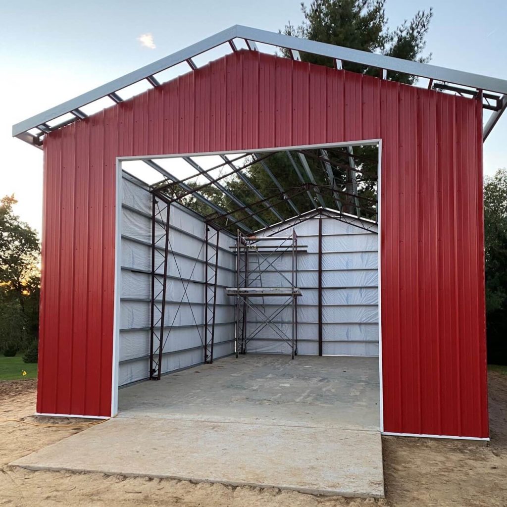 A partially constructed red metal building with an open front, showing an interior frame. The building stands on a concrete slab surrounded by bare ground and some trees in the background.