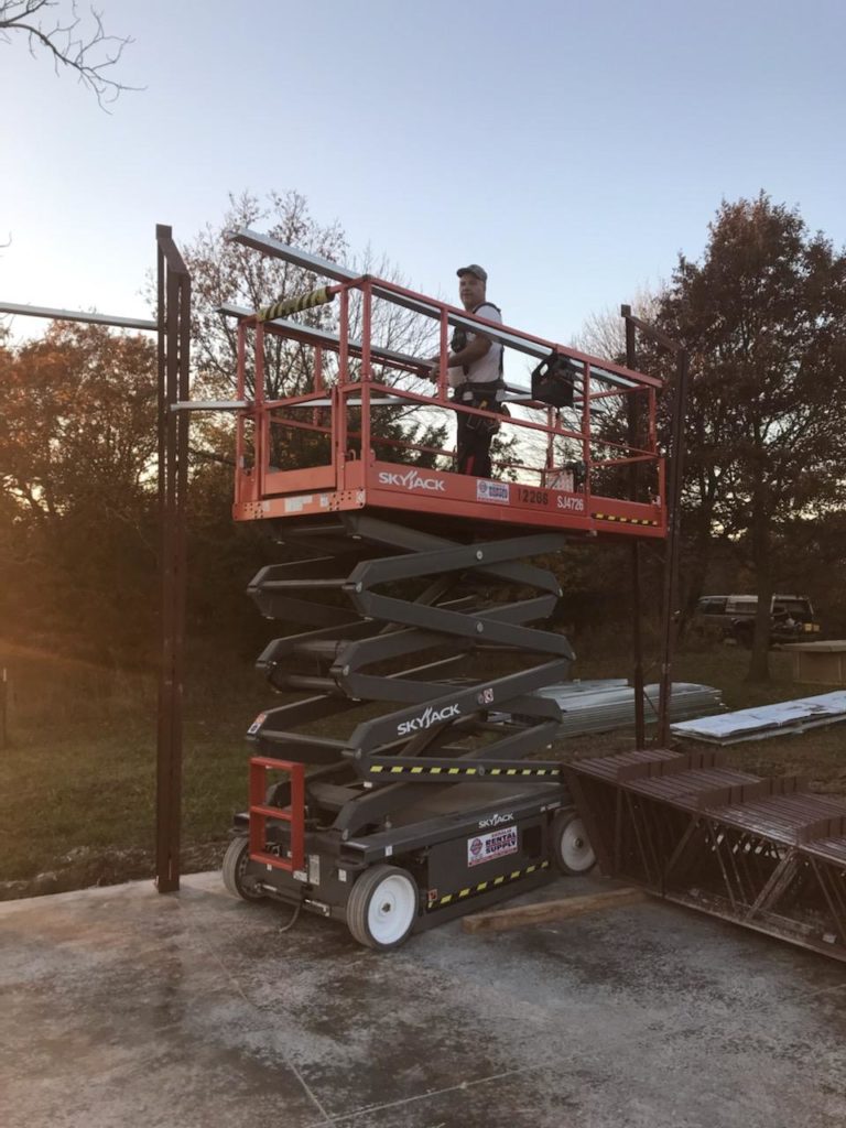 A person stands on a raised scissor lift, working on a metal structure. The lift is labeled "Skyjack." The scene is outdoors with trees in the background and a clear sky.