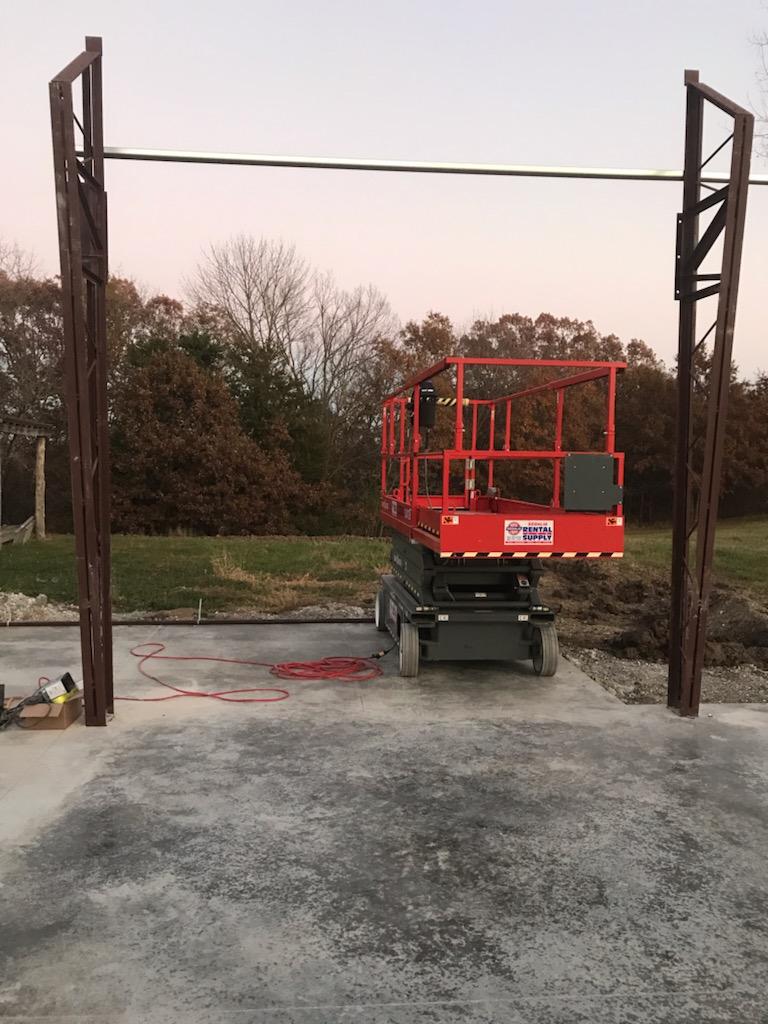 A red scissor lift positioned between two tall metal beams at a construction site. The ground is paved with concrete, and trees are visible in the background under a clear sky.