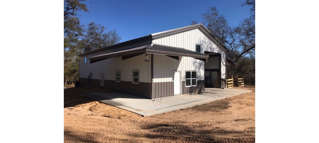 The newly constructed Summerdale Storage is a large metal-sided building with a high roof and several small windows. It features a concrete patio area and is surrounded by dirt and trees, all basking under a clear blue sky.