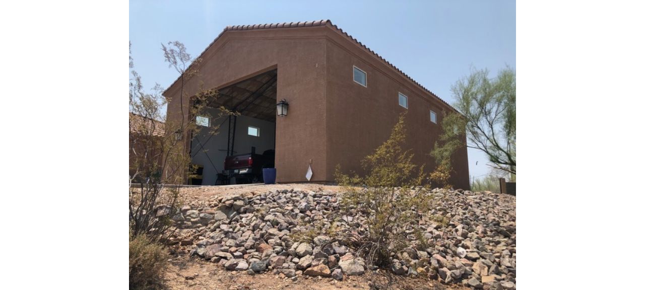 A brown stucco building on Breezy Court features a large open garage door, nestled on rocky, sloped terrain. The sky is clear, and a tree stands to the right. Within the shop's garage, various items and a vehicle are partially visible.