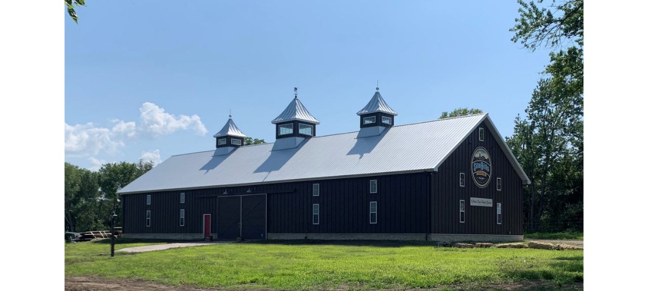 A large, dark brown barn with a metal roof sits amid the verdant fields of Silo Hills Farm on a sunny day. It features three cupolas and a painted logo on the side. Majestic trees surround the structure, and the sky is clear with just a few clouds.