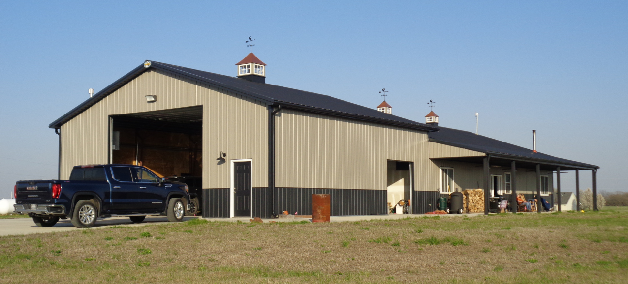 A spacious metal garage, reminiscent of a barndominium, features a black roof with cupolas on top. A black pickup truck sits in front of the open door, with barrels and various items nearby. The sky is clear and the green grass adds to this perfect retirement setting.
