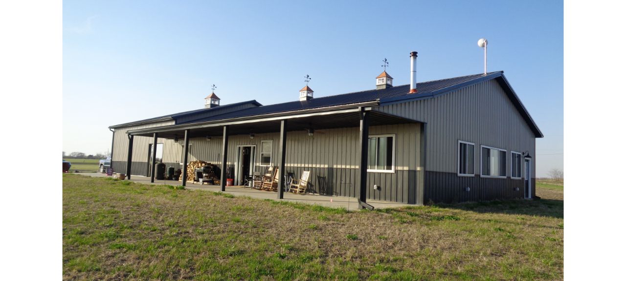 A large gray metal barndominium features a barn-style design with a covered porch, complete with rocking chairs and wooden tables. The roof boasts cupolas and a chimney. A grassy field surrounds this perfect retirement retreat, set against a clear blue sky.
