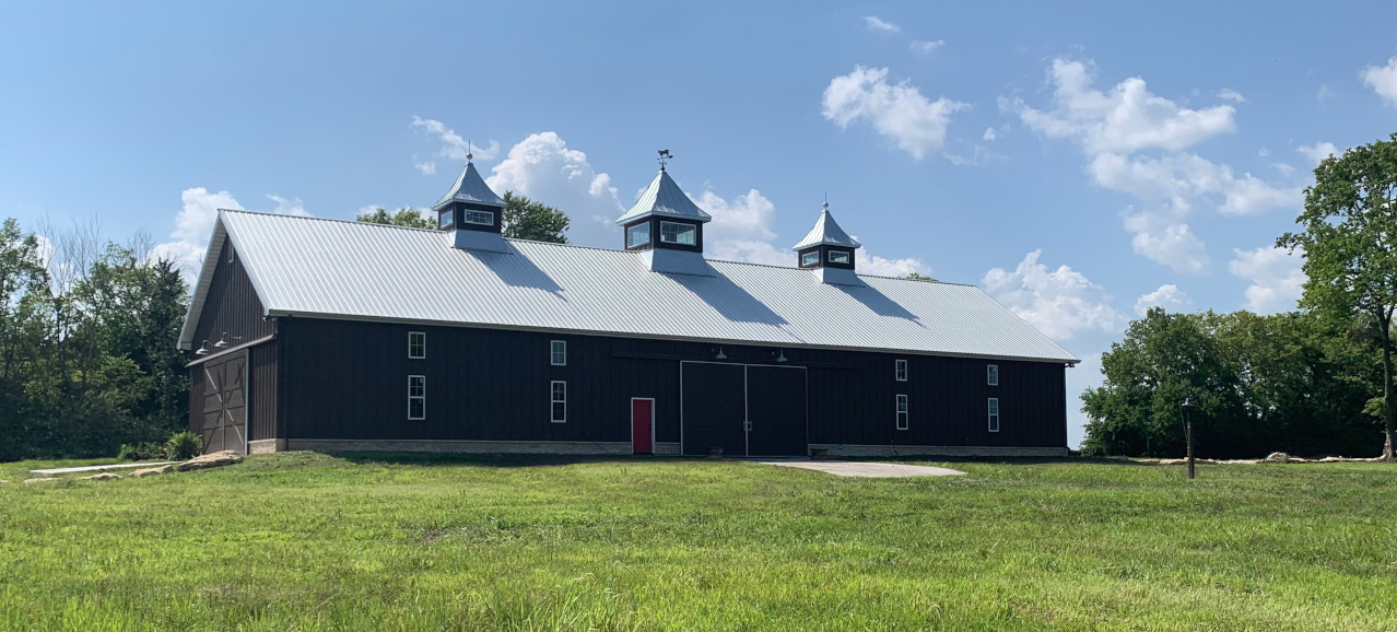 A large black barn with a silver roof and three cupolas stands proudly in the open grassy fields of Silo Hills Farm under a clear blue sky. Trees border the left and right sides of the barn, creating a picturesque country setting.