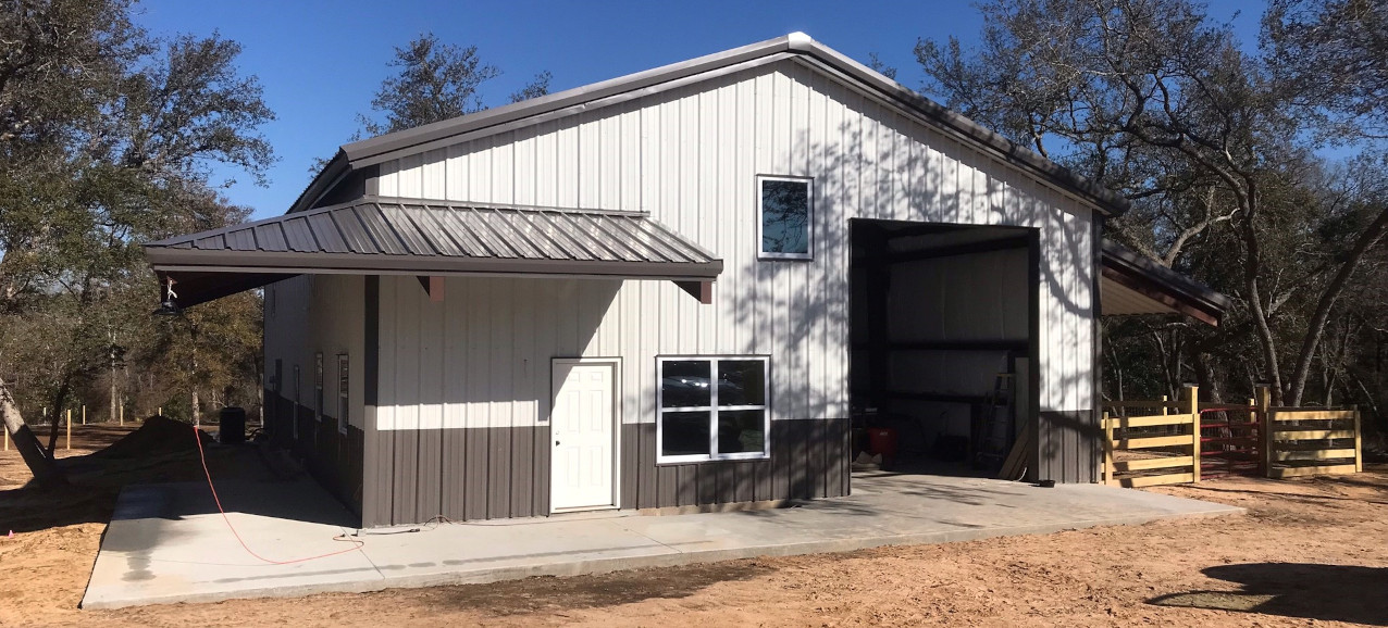 In a dirt clearing surrounded by trees in Santa Clara, a large, newly constructed barndominium with a sloped roof and a white and gray exterior stands. It features a small door, several windows, and an open garage space on the side.