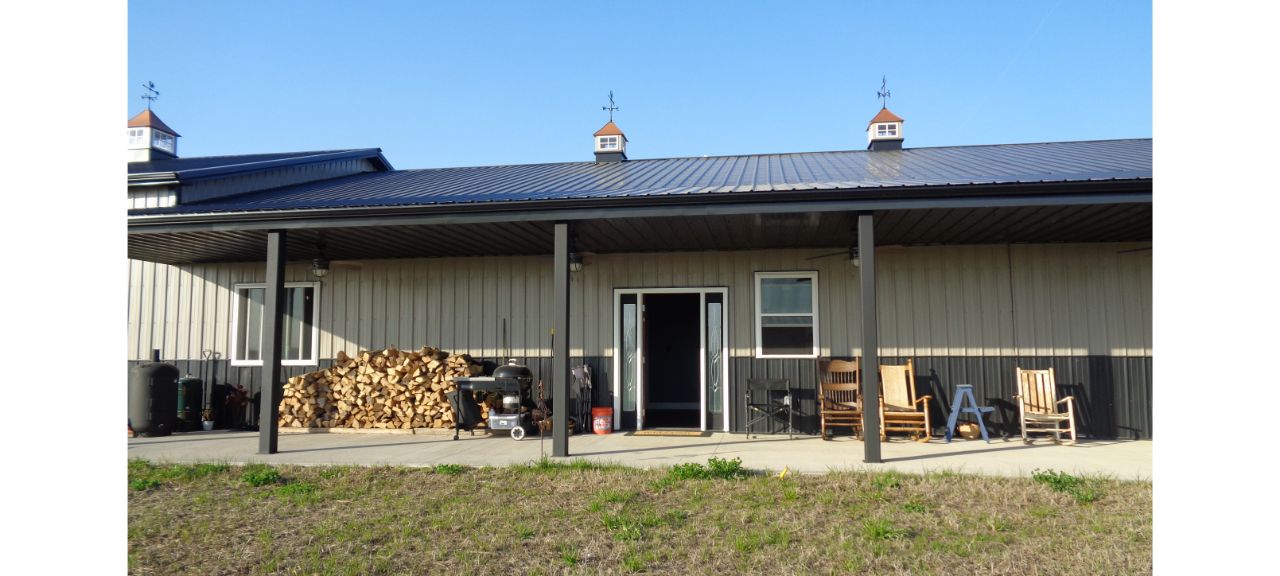 A charming barndominium with a welcoming porch features a stack of firewood, a grill, and rocking chairs, perfect for retirement relaxation. The roof boasts three cupolas, while a grassy area stretches out in the foreground beneath the clear blue sky.