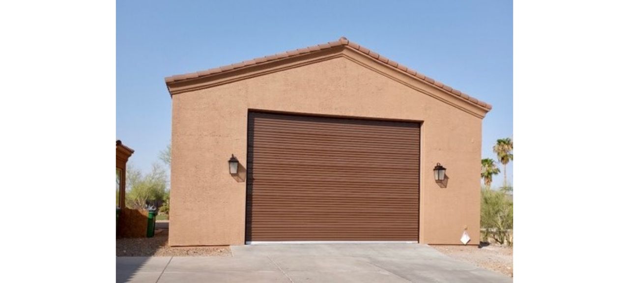 The tan stucco garage on Breezy Court features a dark brown roll-up door, flanked by two wall-mounted lanterns. Under the clear blue sky, palm trees sway gently in the background, adding a serene touch to this picturesque Garage Shop setting.