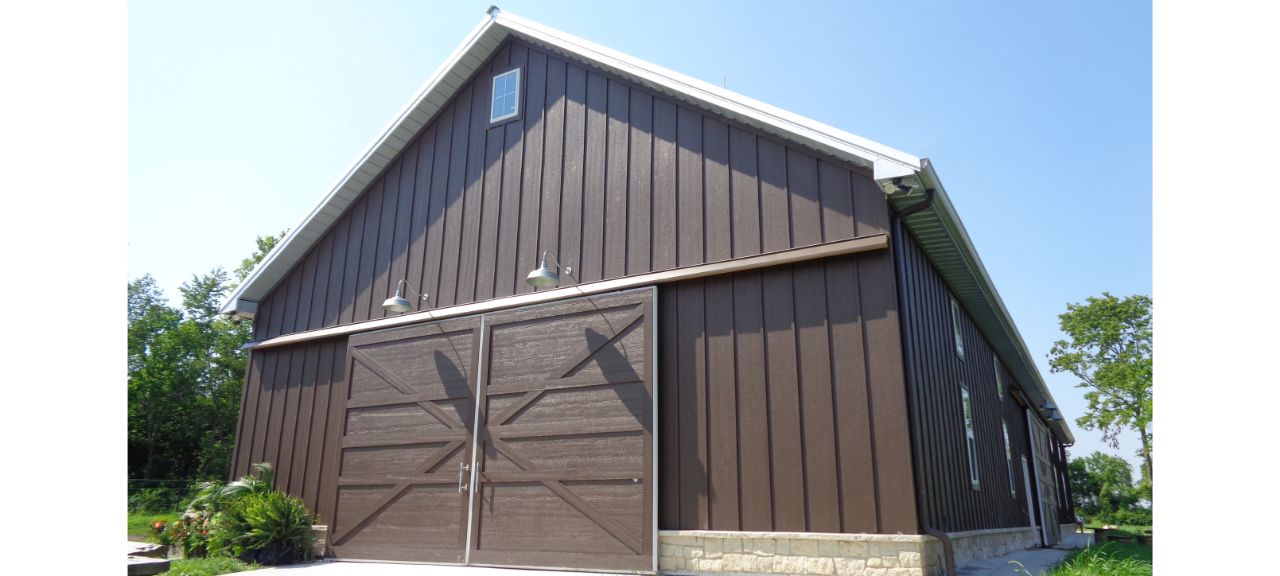 A large brown barn with vertical siding and two wooden double doors stands proudly under the clear blue sky at Silo Hills Farm. The white roof gleams in the sunlight, with two outdoor lights above the doors. Lush green foliage can be seen to the left and in the background.