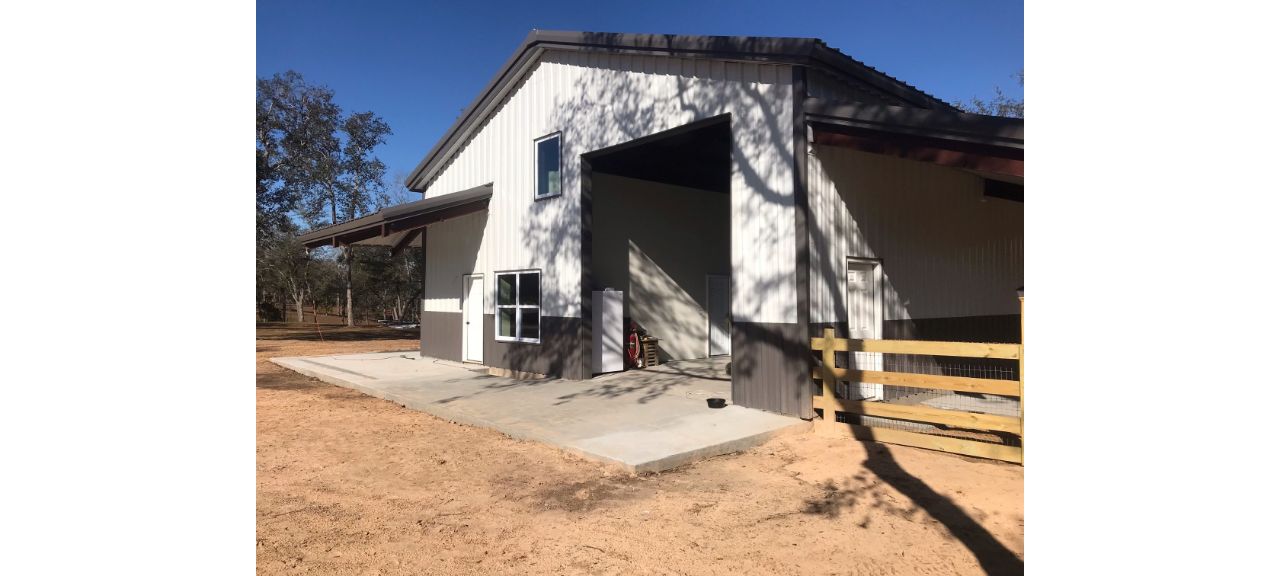 A large, open barn with a high roof stands on a dirt and concrete base, reminiscent of the designs at Summerdale Storage. An open sliding door, side door, and window invite a gentle breeze. A wooden fence is visible on the right, while trees cast shadows across this charming storage facility under a clear blue sky.