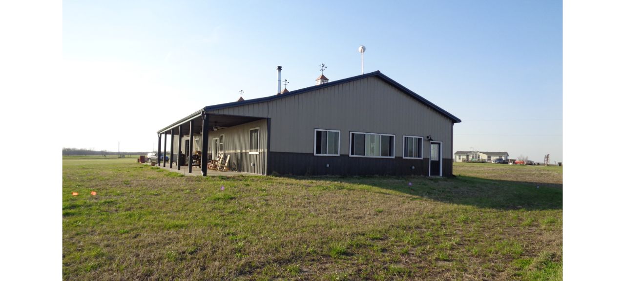 A single-story, metal-sided barndominium with a sloped roof stands on a grassy field. The building has a porch with outdoor seating and several windows, perfect for relaxation in retirement. The sky is clear and blue.
