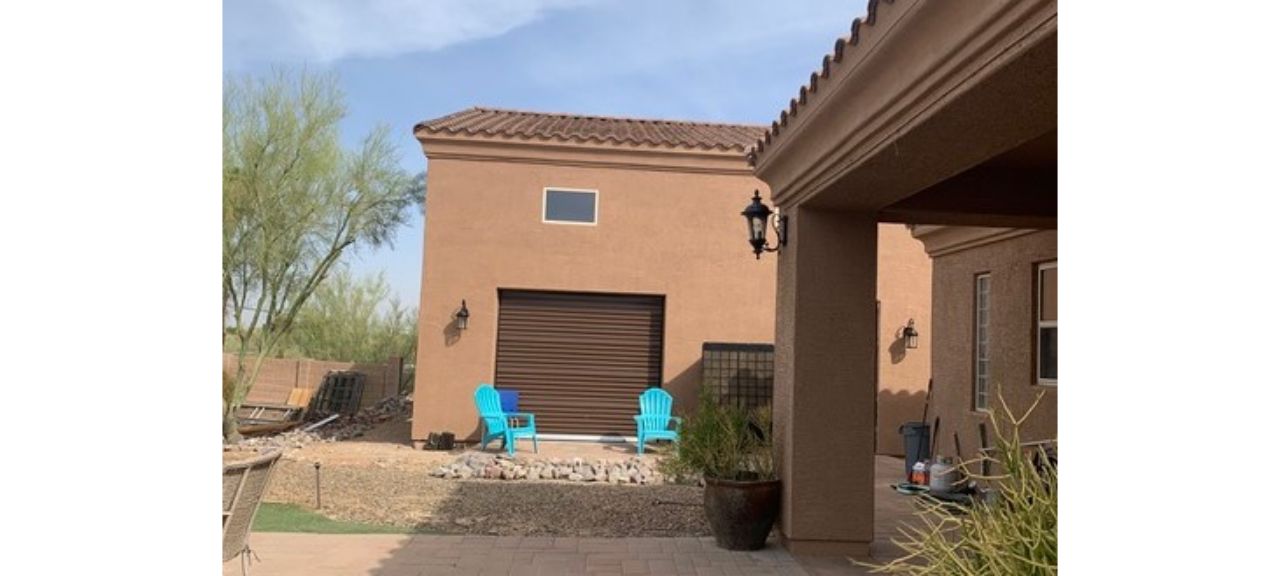 Two blue chairs sit on the patio at Breezy Court, facing a brown building with a closed garage door, reminiscent of a quaint garage shop. A light fixture adorns the wall as desert plants flourish in the background under a clear sky.