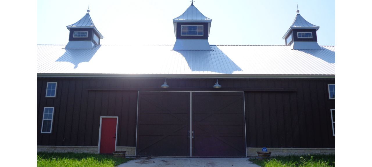 A large barn at Silo Hills Farm features a dark wooden exterior with three cupolas on its metal roof. Two large sliding doors and a smaller red door to the left add charm, as sunlight casts shadows on the structure. Grass grows along the base, blending seamlessly into the surrounding hills.
