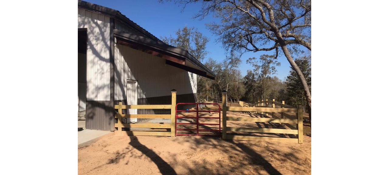 A red metal gate is attached to a wooden fence in front of the barn belonging to Summerdale Storage, offering practical storage solutions. Trees and a clear blue sky paint a serene backdrop, while shadows dance on the ground and the building’s slanted roof.