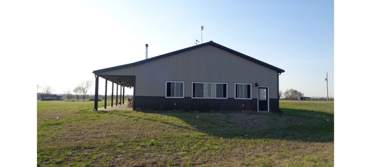 A cozy barndominium with a metal roof sits in an open grassy field, perfect for a peaceful retirement. It features several windows, a front door, and a shaded porch on one side. The sky is clear and blue.
