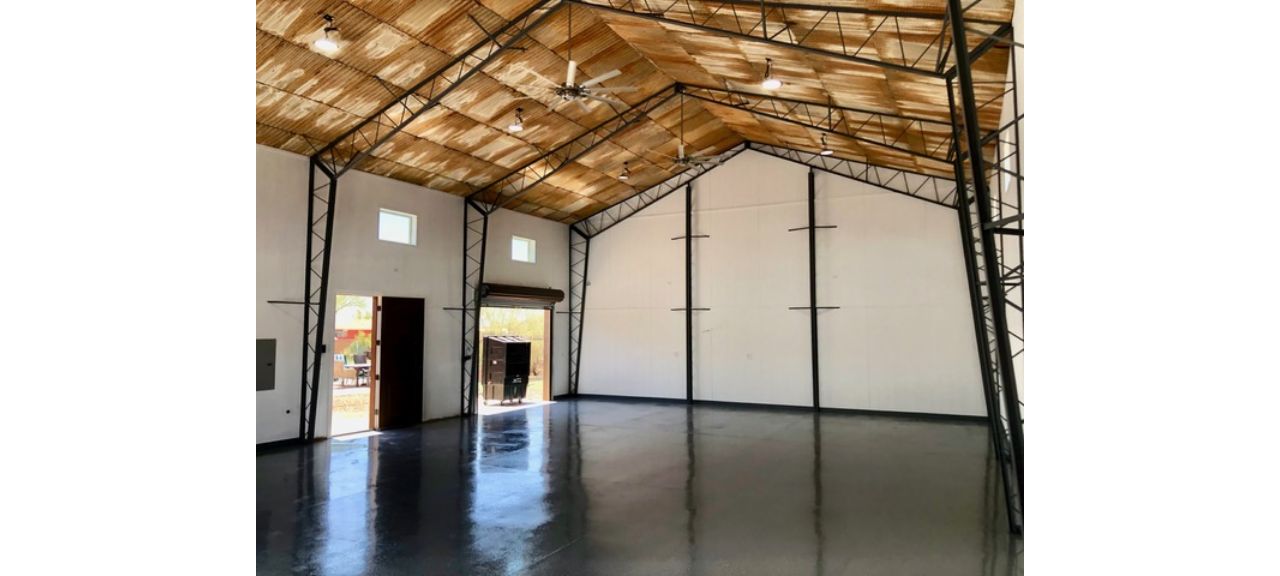 A spacious, empty warehouse interior with a shiny concrete floor and high vaulted ceiling resembles a versatile shop. The white walls with exposed black steel beams lend an industrial charm. Natural light breezes in through small upper windows and a partially open entrance, enhancing its airy vibe.