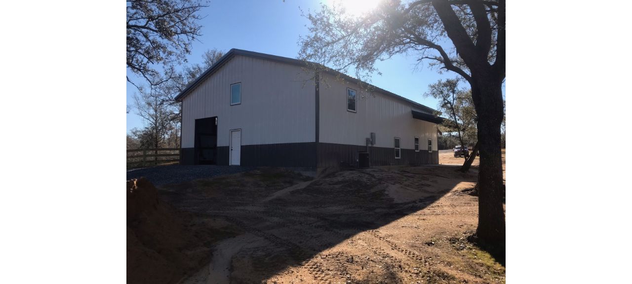 A large, light-colored barn with a dark roof stands proudly under a clear blue sky at Summerdale Storage. The structure is surrounded by leafless trees, sunlight streaming through the branches, casting shadows on the sandy ground.
