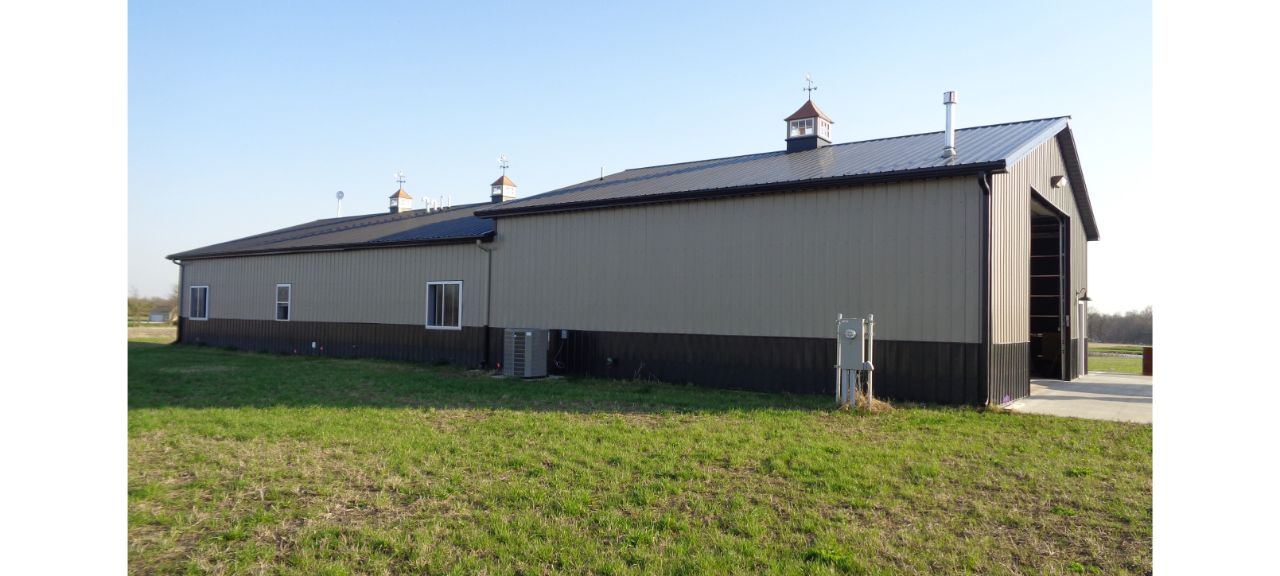 A spacious metal barndominium with a black roof and beige walls stands proudly on a grassy field. Cupolas adorn the rooftop, while several windows line the sides. Perfect for retirement, a concrete path leads invitingly to the large open doorway at the front.