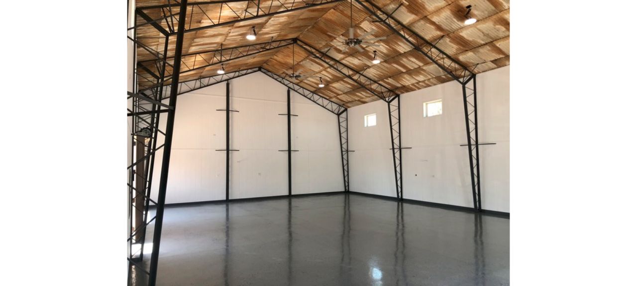 Spacious empty warehouse interior with polished concrete floors, white walls, and a high wooden ceiling supported by metal beams. Several small windows allow natural light to flow in like a breezy court, while multiple overhead lights are installed. Perfect for transforming into a unique shop or garage space.