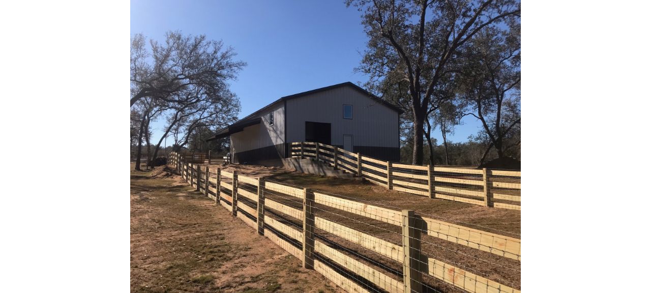 A gray barn in a rural setting with a dark roof stands proudly as the perfect Summerdale Storage solution. Surrounded by a wooden fence, with a few bare trees nearby, the sky is clear and blue, suggesting a bright, sunny day.