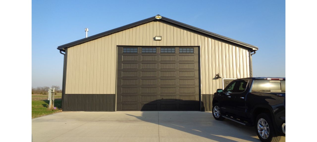A large beige and black metal barndominium with a high, closed double door stands proudly. The building rests on a concrete driveway with a black truck parked in front, ready for the adventures of retirement against the backdrop of a clear blue sky.
